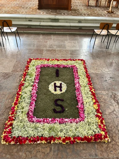 Blumenteppich aus Rosenblättern, Gräsern und weißen Blüten, in der Mitte das Christusmonogramm IHS