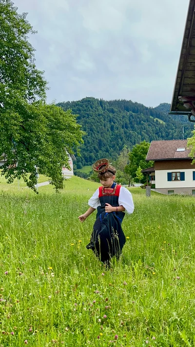 Ein Mädchen in Bregenzerwälder Tracht mit goldenem Kopfschmuck geht durch eine hohe Blumenwiese